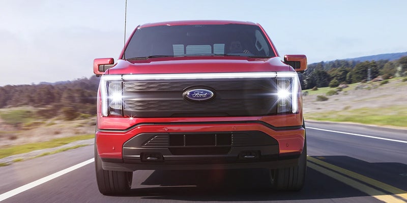 Front view of a red 2022 Ford F-150 Lightning with the headlights on being driven on the highway. | Ford dealer in Little Rock, AR.